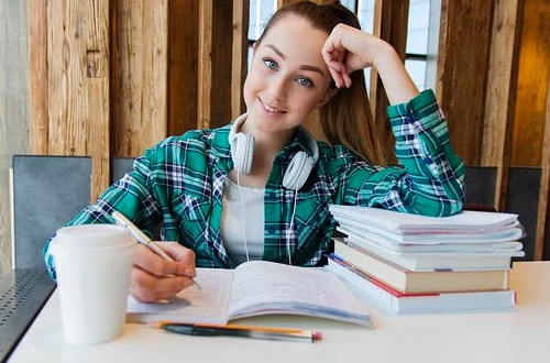Smiling girl takes notes in a notebook. There is a pile of books and a cup on the desk.